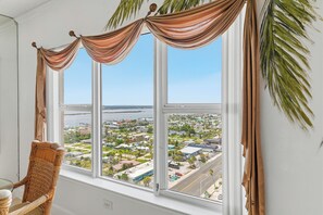 A room with a large window overlooking a residential area, a wicker chair, and a view of the water. - 🌴Panoramic Ocean + River Views 19th Floor Corner Condo - Towers Ten 1901 (Daytona Beach Shores)