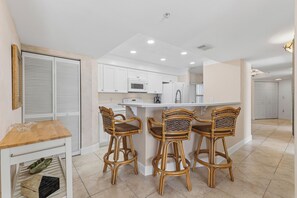 A kitchen with white cabinets, a microwave, and a refrigerator. There are three wicker chairs at a small island. - 🌴Panoramic Ocean + River Views 19th Floor Corner Condo - Towers Ten 1901 (Daytona Beach Shores)