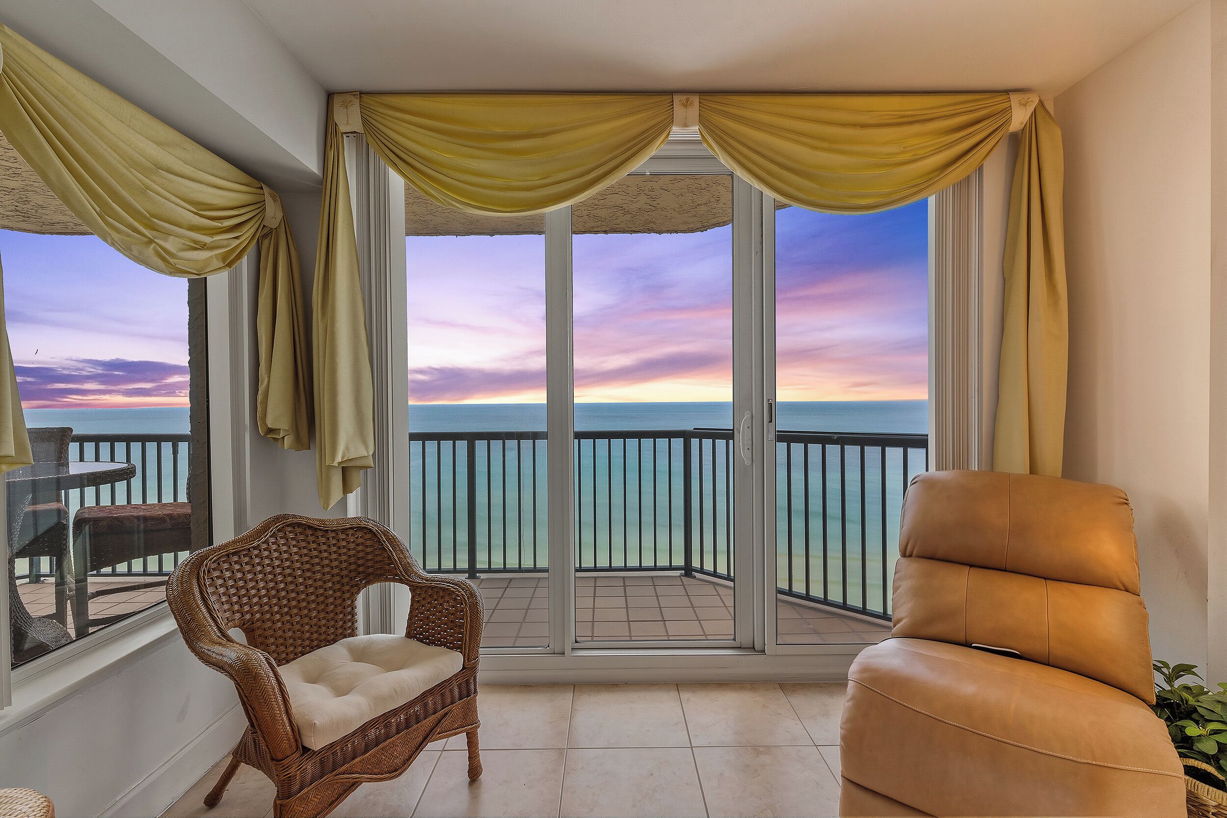A balcony with a wicker chair and a leather sofa, overlooking a body of water with a sunset view.