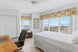 A bedroom with a bed, a chair, a desk, a ceiling fan, and a view of a cityscape through large windows. - 🌴Panoramic Ocean + River Views 19th Floor Corner Condo - Towers Ten 1901 (Daytona Beach Shores)