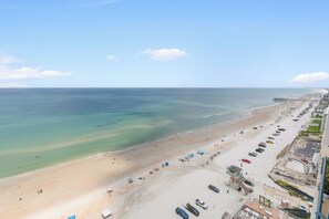 A beach with a pier, clear skies, and a parking area with vehicles. - 🌴Panoramic Ocean + River Views 19th Floor Corner Condo - Towers Ten 1901 (Daytona Beach Shores)