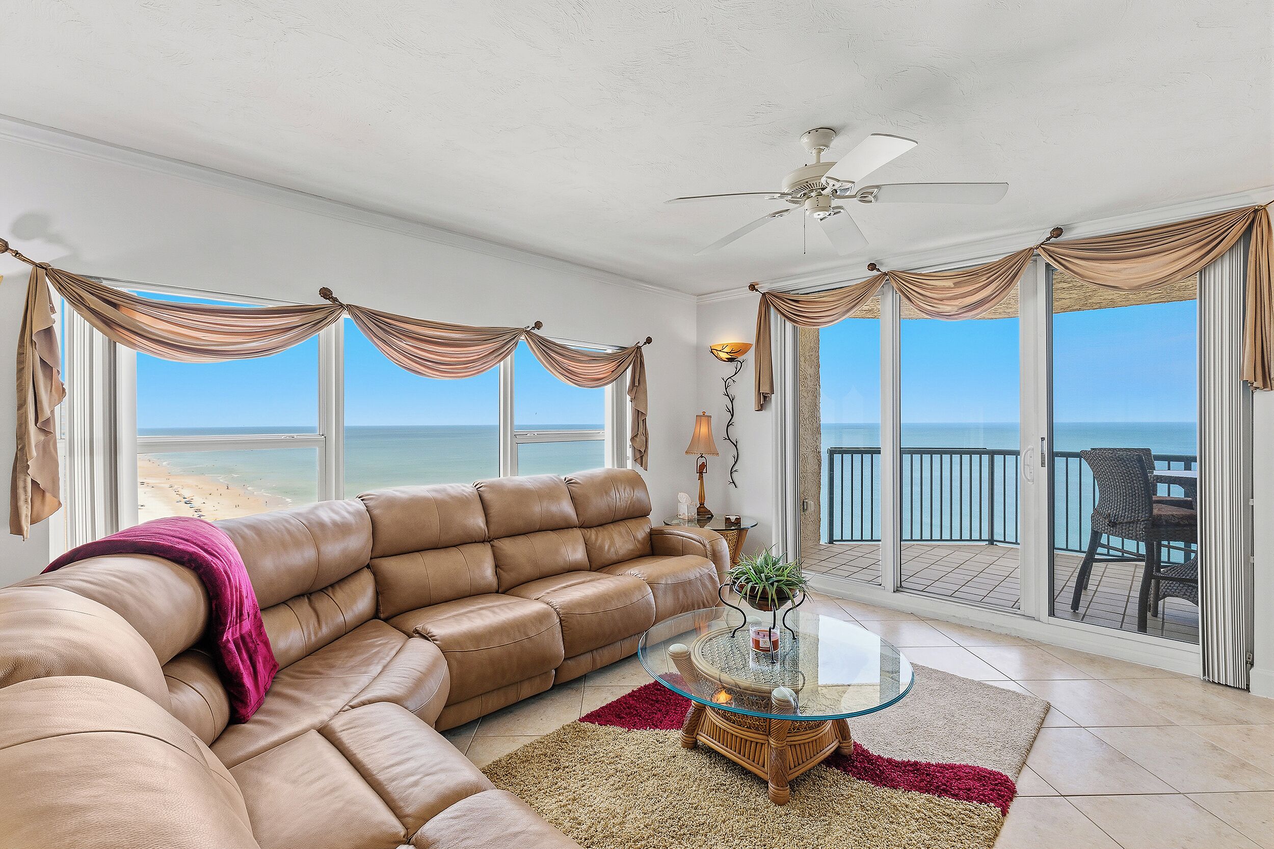 A living room with a large leather sofa, a glass coffee table, and a view of the beach.