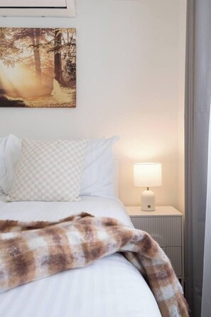 Une chambre à coucher avec un lit, un coussin à carreaux, une couverture, une lampe de chevet et un tableau représentant une forêt, accroché au mur.