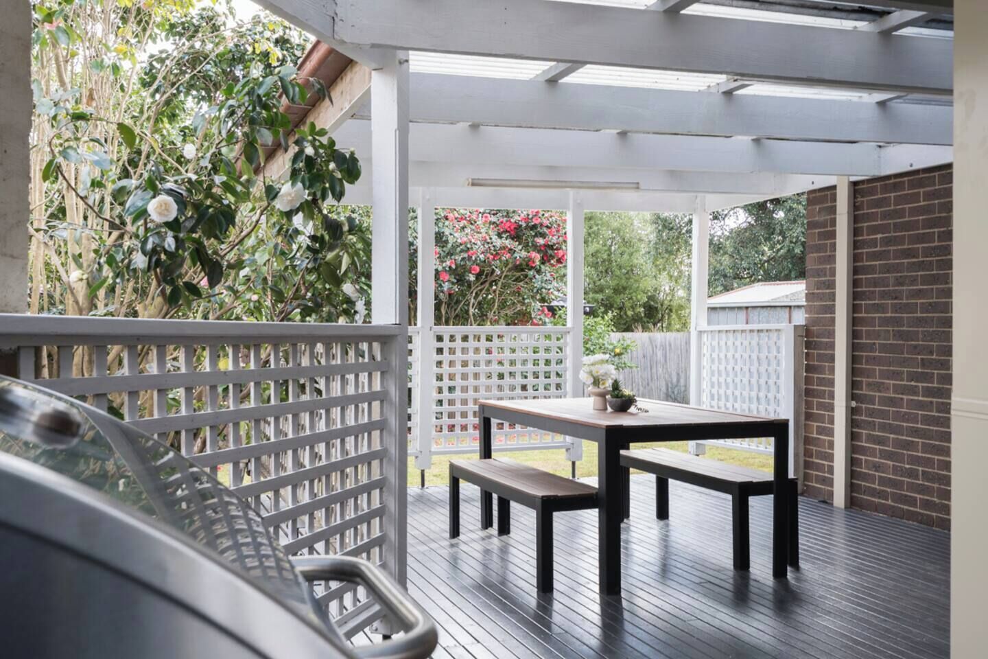 A covered patio area with a wooden table and benches, surrounded by a white fence and lush greenery.