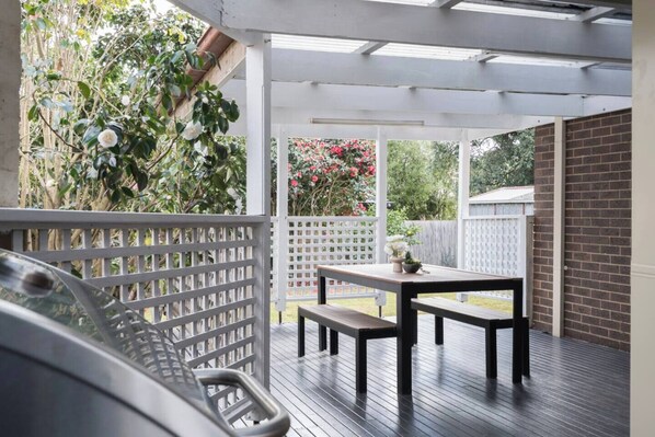 Un patio cubierto con una mesa de madera y bancos, rodeado por una cerca blanca y abundante vegetación.