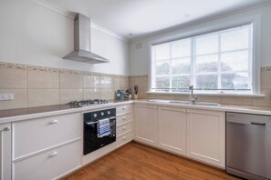 Una cocina con estufa a gas, horno y lavavajillas, una ventana con persianas y salpicadero de azulejos.