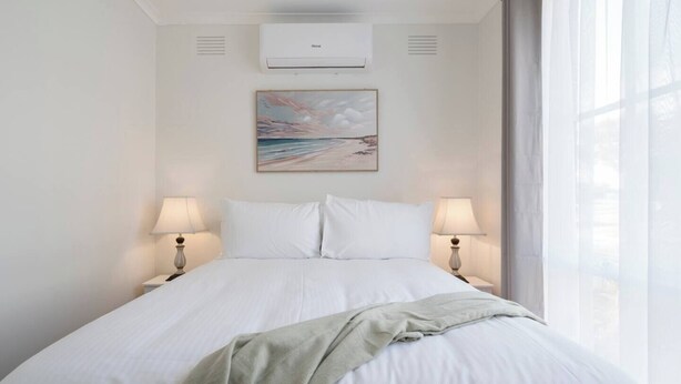 Una camera d'hotel con un letto, due lampade ai lati del letto, un condizionatore, un quadro appeso al muro e una finestra con tende.