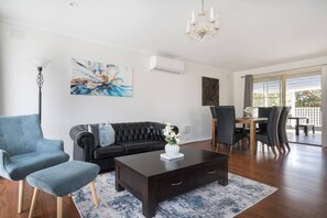 Un salon moderne avec un canapé marron foncé, un fauteuil bleu et une table basse sur laquelle est posé un vase. On y trouve également un coin repas avec une table et des chaises, ainsi qu’une grande fenêtre offrant une vue dégagée.