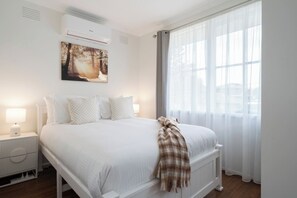 Una habitación de hotel con cama, mesita de noche, aire acondicionado mural, ventana con cortinas traslúcidas y un cuadro sobre la cama.