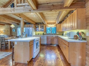 A wooden cabin interior with a kitchen featuring wooden cabinets, a central island, and a dining area with chairs.