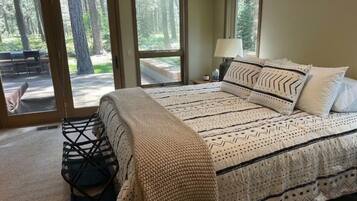 A bedroom with a large bed, a nightstand, a lamp, and a view of a forest through sliding glass doors.