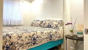 Un dormitorio con cama, ventana con cortinas, mesita de noche con lámpara y un florero con flores.