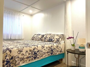 A bedroom with a bed, a window with curtains, a bedside table with a lamp, and a vase with flowers.