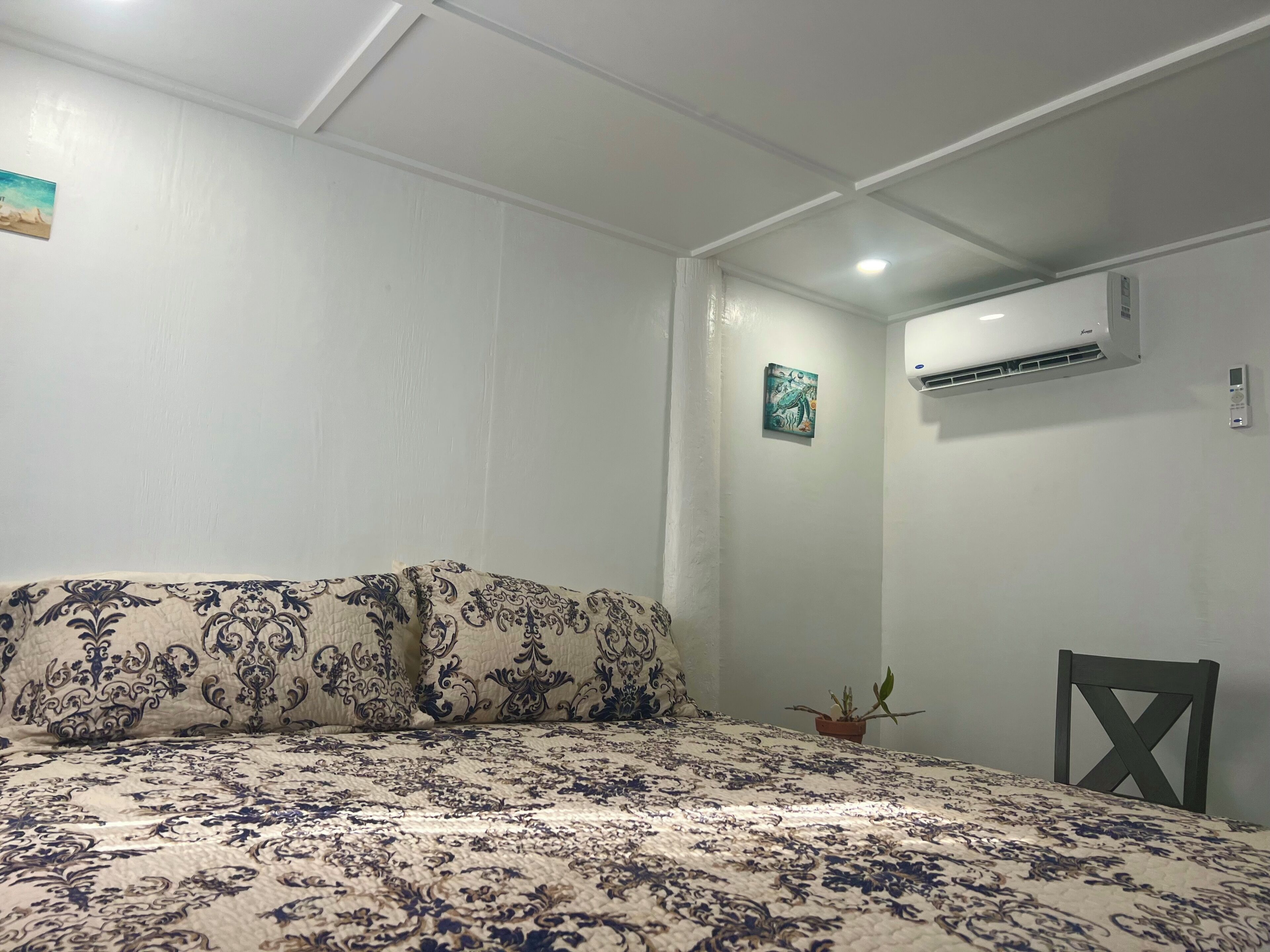 A bedroom with a bed, a potted plant, an air conditioning unit, and a wall-mounted artwork.