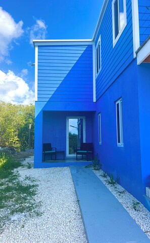 A modern house with a blue exterior, a covered patio with seating, and a gravel pathway.