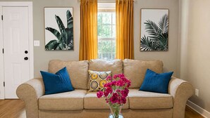 Een woonkamer met een beige bank, blauwe kussens, een vaas met roze bloemen en twee kunstwerken met plantenmotieven aan de muur.