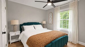 Een slaapkamer met een bed, twee nachtkastjes, een plafondventilator, een raam met gordijnen en uitzicht op groen.