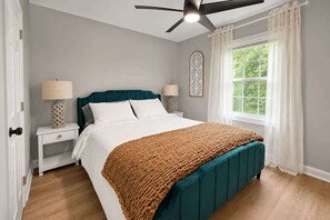 A bedroom with a bed, two bedside tables, a ceiling fan, a window with curtains, and a view of greenery outside. - Downtown Duplex with Fenced Yard (Raleigh)