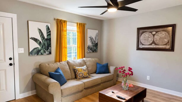 Un salon avec un canapé beige, une table basse, un ventilateur de plafond et des œuvres d’art accrochées aux murs.