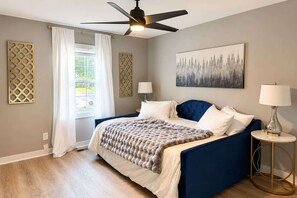 Une chambre à coucher avec un lit, un ventilateur de plafond, une fenêtre avec des rideaux, un tableau au mur et une table de chevet avec une lampe.