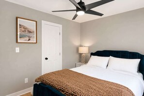 Un dormitorio con cama, ventilador de techo, puerta, mesita de noche con lámpara y un cuadro en la pared.