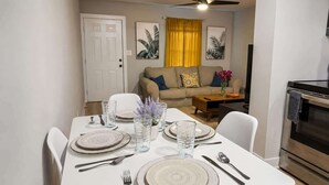 Un coin repas avec une table blanche dressée pour quatre personnes, un canapé en arrière-plan et une cuisine moderne équipée d’une cuisinière.