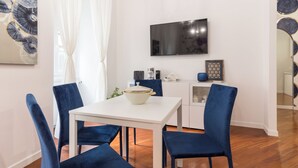 A modern dining area with a white table, blue chairs, a flat-screen TV, and a kitchen area with cabinets and a countertop. - Trastevere | Piazza Trilussa | Rome on Foot—Moro7— (Roma)