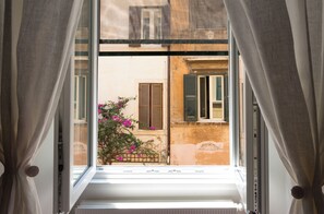 An open window with curtains, a view of a building with shutters, and a flowering plant outside. - Trastevere | Piazza Trilussa | Rome on Foot—Moro7— (Roma)