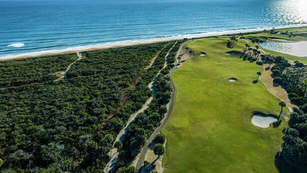 Vista aérea de um campo de golfe, com uma praia e o oceano ao fundo.