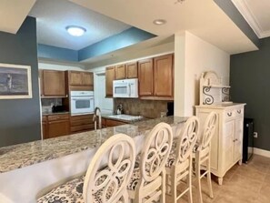 A kitchen with a central island, white bar stools, and wooden cabinets. - Water-View 2BR Condo w/ Pool, Spa & Resort Access (Palm Coast)