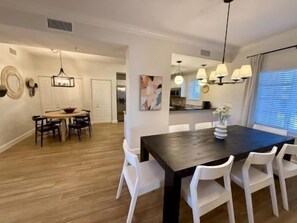 A dining area with a dark wooden table and white chairs, a kitchen in the background, and a large window with blinds. - Bright 3BR Condo w/ Hammock Beach Resort Access (Palm Coast)