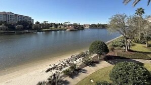 Een meer met een zandstrand, een hotel op de achtergrond, en een verzorgd gazon met bomen.