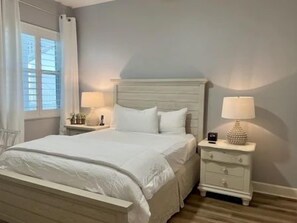 A neatly made bed with white bedding, a bedside table with a lamp, and a window with curtains. - Ocean-View 3BR Condo | Hammock Beach Resort Access (Palm Coast)