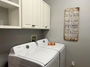 A laundry area with two white front-loading washing machines and a wall-mounted sign with text. - Ocean-View 3BR Condo w/ Pool & Resort Access (Palm Coast)