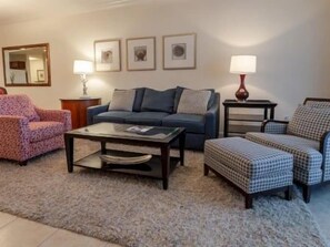 A living room with a sofa, armchairs, a coffee table, and a rug. There are lamps and framed pictures on the walls. - Waterway 2BR Condo | Hammock Beach Resort Access (Palm Coast)