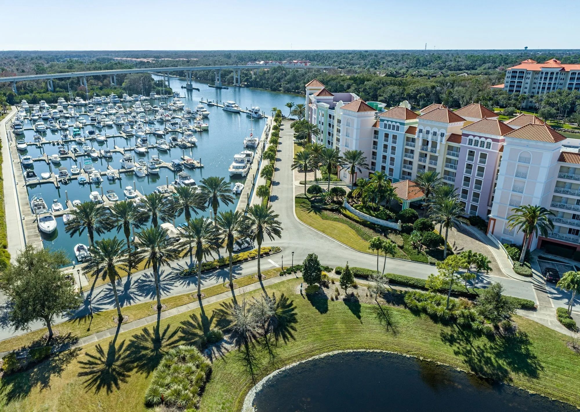 A marina with numerous boats docked, surrounded by a well-maintained landscape with palm trees and a large hotel building with a red roof.