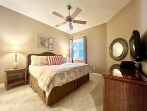Ein Schlafzimmer mit einem Bett, Nachttisch, Lampe, Deckenventilator, Fenster und einem Spiegel auf einer Kommode.
