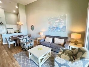 A living room with a sofa, coffee table, and patterned armchair. There is a dining area with a table and chairs. A kitchen is visible in the background. - Oceanside 3BR Condo w/ Hammock Beach Resort Access (Palm Coast)