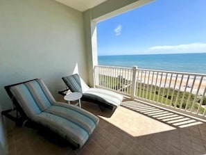 Ein Balkon mit zwei Liegestühlen und einem kleinen Tisch, mit Blick auf einen Strand und den Ozean.