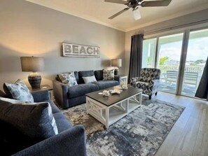 A living room with a sofa, armchair, coffee table, and a 'BEACH' sign on the wall. - Lakeside 3BR Condo | Balcony, Pool + Resort Access (Palm Coast)