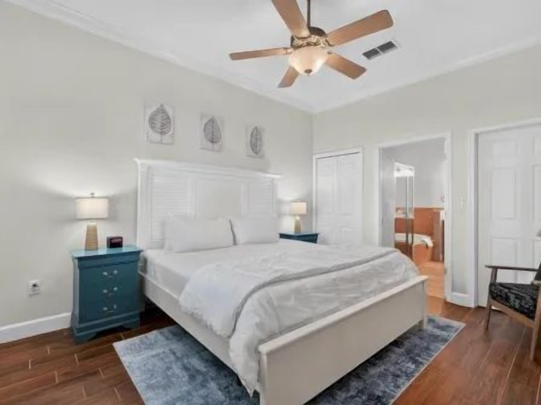 Un dormitorio con cama, mesitas de noche, ventilador de techo y una puerta que da a otra habitación.