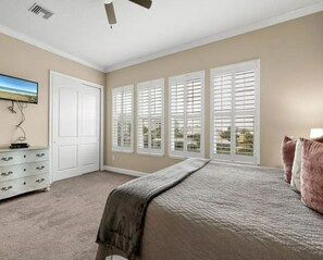 A bedroom with a bed, a dresser, a television, and a window with shutters. - Lake-View 11BR House | Heated Pool & Resort Access (Palm Coast)