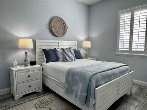 A bedroom with a bed, two bedside tables, a nightstand, a wall clock, a window with blinds, and a decorative wall piece. - Lake-View 5BR Home w/ Hammock Beach Resort Access (Palm Coast)