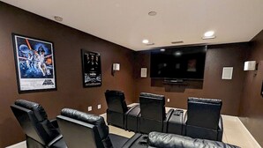 Sala de cine en casa con butacas reclinables de cuero negro, un televisor de pantalla plana grande y carteles de películas en la pared.