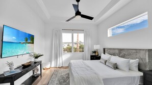 Ein Schlafzimmer mit Bett, Deckenventilator, Fernseher, Fenster mit Vorhängen und Blick auf den Strand.