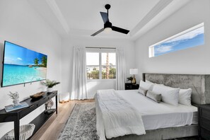 Ein Schlafzimmer mit Bett, Deckenventilator, Fernseher, Fenster mit Vorhängen und Blick auf den Strand.