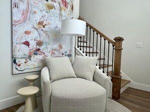 Ein Zimmer mit einer Treppe, einem großen abstrakten Gemälde, einem weißen Sessel und einem kleinen Hocker.