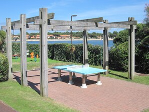 A ping pong table under a wooden pergola with a view of a beach and buildings in the background. - T2 face au Lac | À 100 m de la plage | Piscine (Vieux-Boucau-les-Bains)