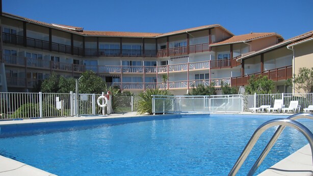Una piscina con una escalera metálica, rodeada por una valla blanca, y un edificio con balcones al fondo.