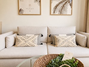 Ein sofasitz in neutraler Farbe, mit gemusterten Zierkissen und einem gewebten Korb mit Grünpflanzen und einer Karte auf einem Glastisch.